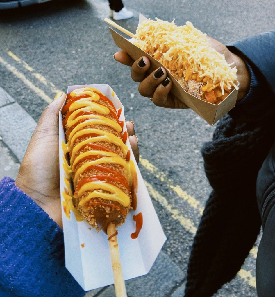 two corndogs in hands. one coated in ketchup and mustard. the other with a pile of grated cheese over top.